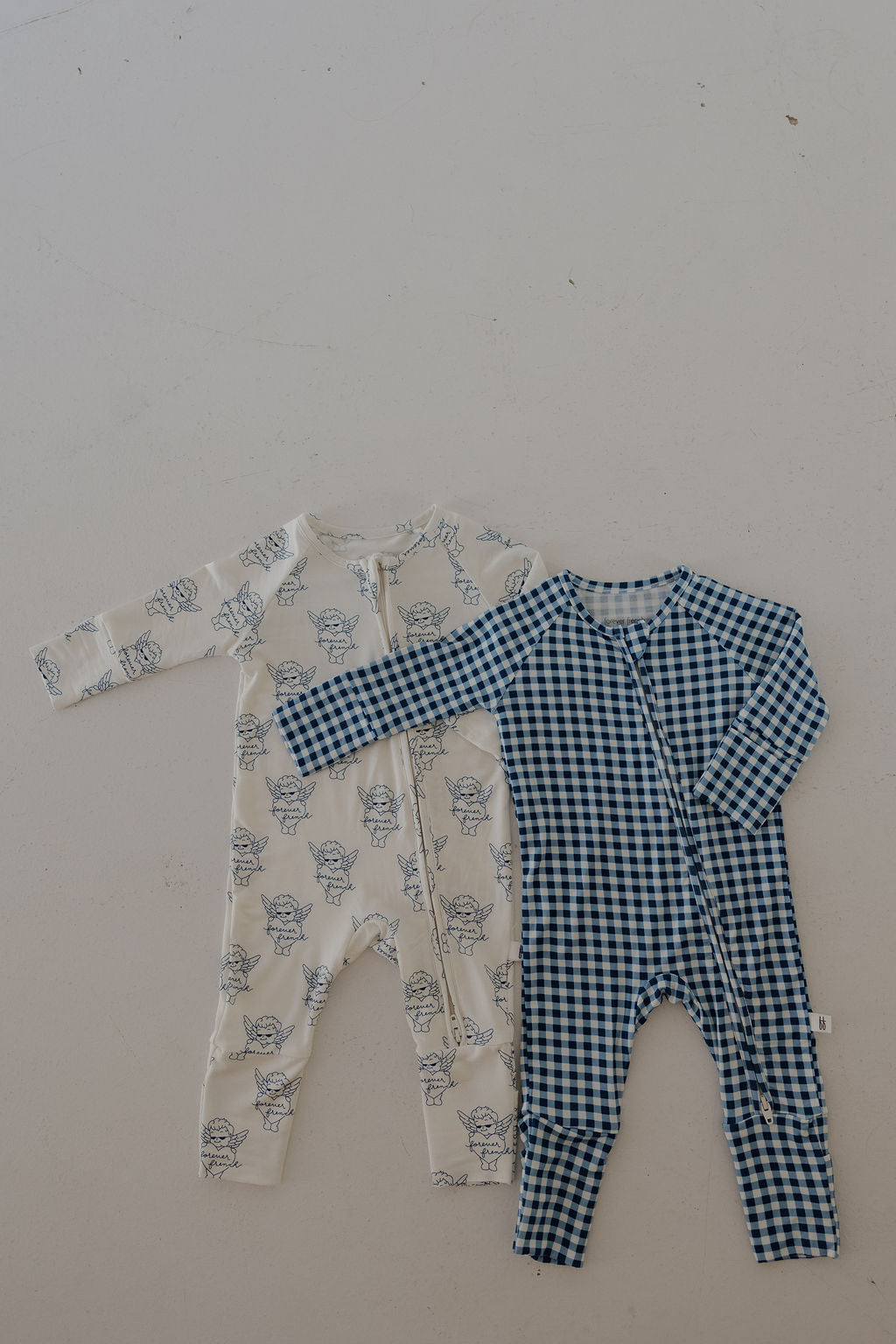 Two forever french baby Bamboo Zip Pajamas | Blue Gingham sleepers are displayed: one features a white base with a blue bear face, the other has a blue gingham pattern. Both have front zippers and are made from breathable baby sleepwear fabric.