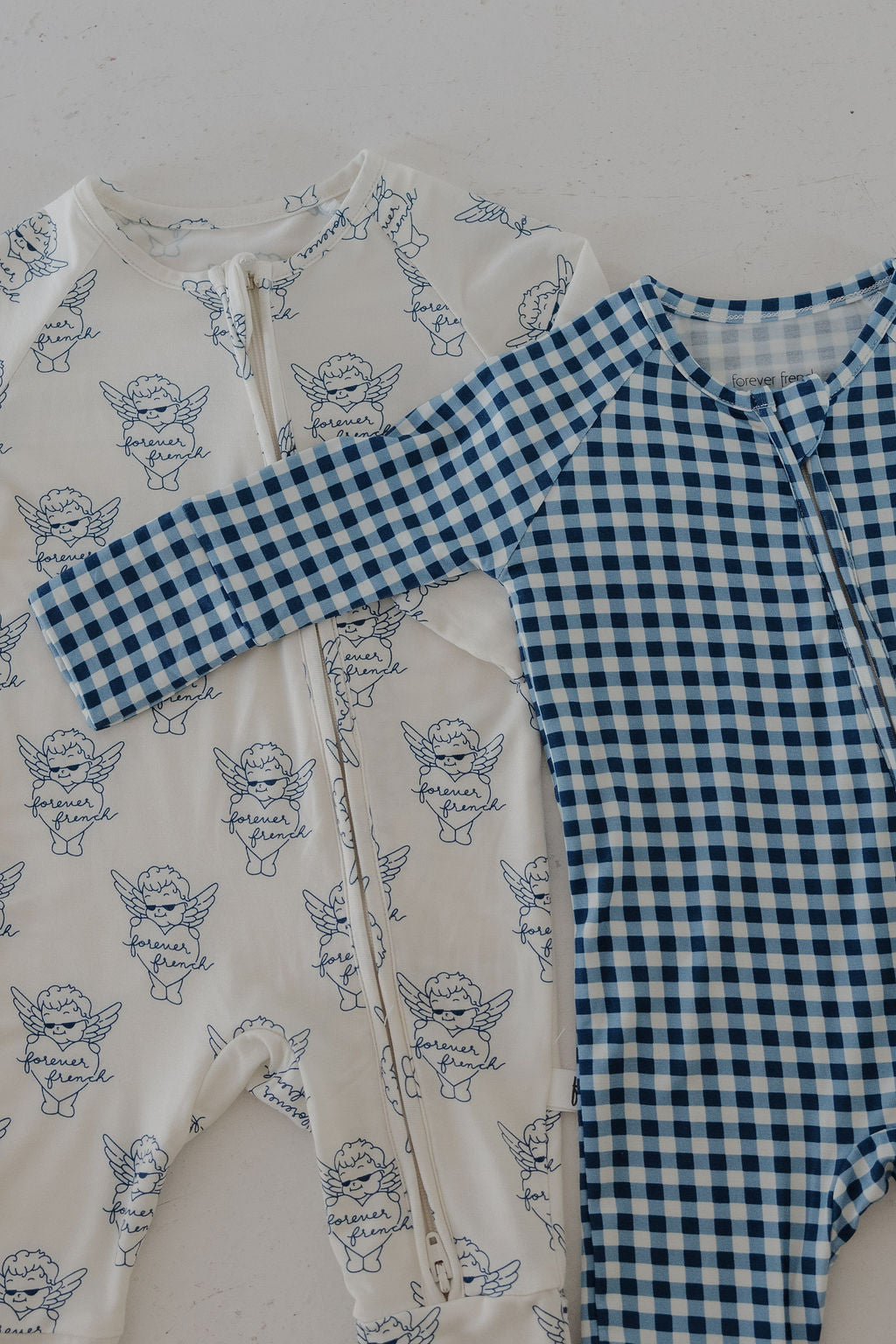 Two forever french baby Bamboo Zip Pajamas lay side by side: one in blue gingham checks, the other white with a blue angel print. Both offer breathable sleepwear with front zippers and long sleeves for all-night comfort.