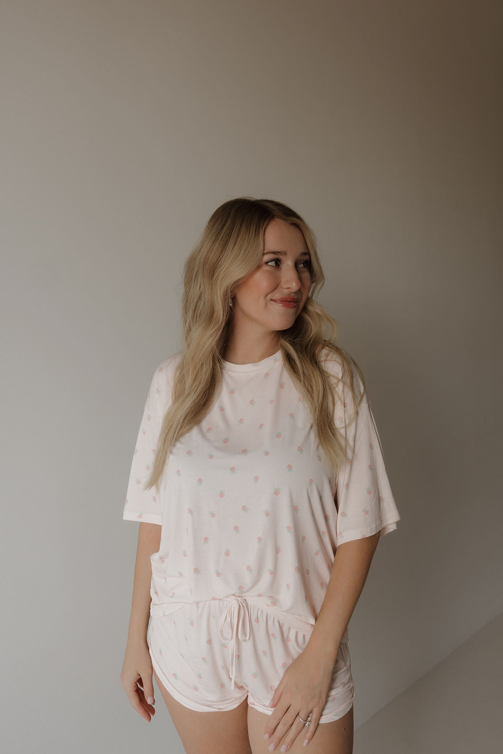 A woman with long blonde hair smiles and looks slightly to her left while wearing the forever french baby Women’s Bamboo Short Set | Rosie—a light pink, breathable pajama set with small dots—standing against a plain, neutral wall.