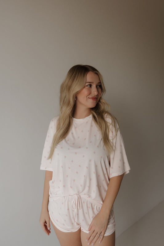 A woman with long blonde hair smiles and looks slightly to her left while wearing the forever french baby Women’s Bamboo Short Set | Rosie—a light pink, breathable pajama set with small dots—standing against a plain, neutral wall.