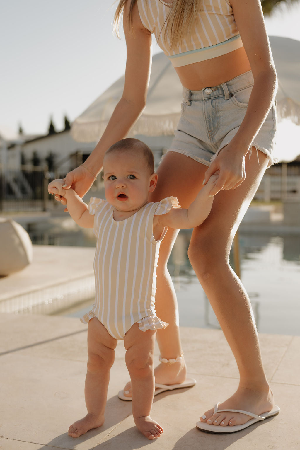 A baby wearing the lolo webb 2 Girl Sleeveless Swimsuit | Sun Rays, featuring yellow and white stripes with ruffles, stands by a pool holding hands with a woman; sunny scene with umbrellas and outdoor furniture in the background.