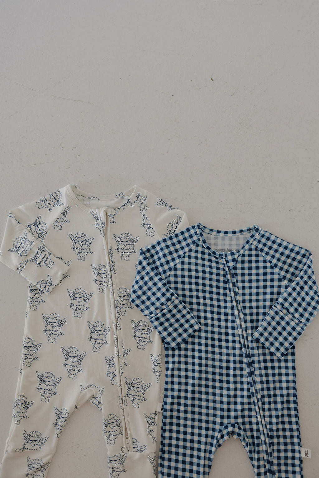 Two forever french baby Bamboo Zip Pajamas are laid flat: one features blue cherub prints on white, and the other showcases a blue gingham pattern. Both are breathable zip-up sleepwear, perfect for cozy nights.