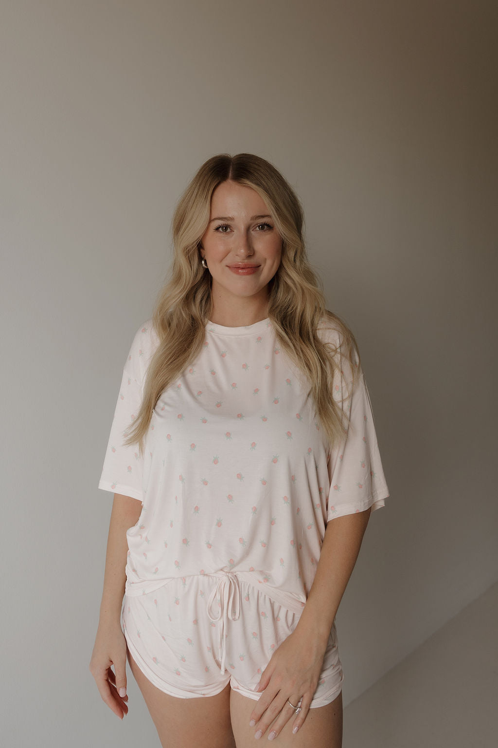 A woman with long blonde hair is smiling at the camera while wearing the forever french baby Women’s Bamboo Short Set | Rosie—a breathable, hypoallergenic bamboo pajama set in light pink with subtle patterns and short sleeves—standing against a plain, light background.