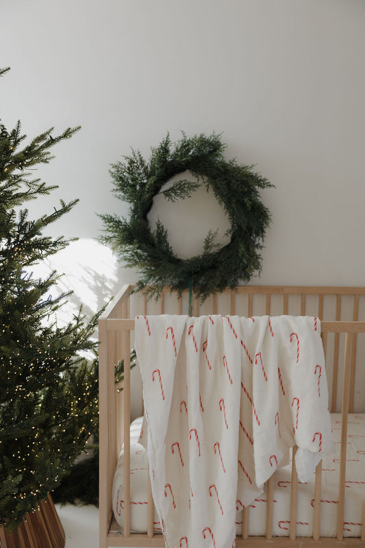 A forever french baby Muslin Quilt | Candy Cane (Preview) in white with red candy canes is draped over a wooden crib with nursery bedding. A green wreath hangs behind, and a lit Christmas tree stands nearby.