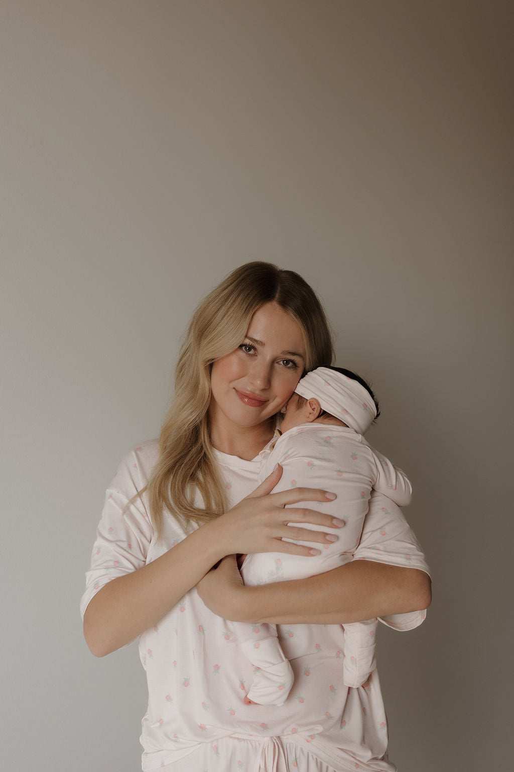 A woman with long blonde hair, dressed in the forever french baby Women’s Bamboo Short Set | Rosie in light pink, smiles gently while holding a swaddled baby wearing matching breathable and hypo-allergenic pink clothes and headband, set against a neutral background.