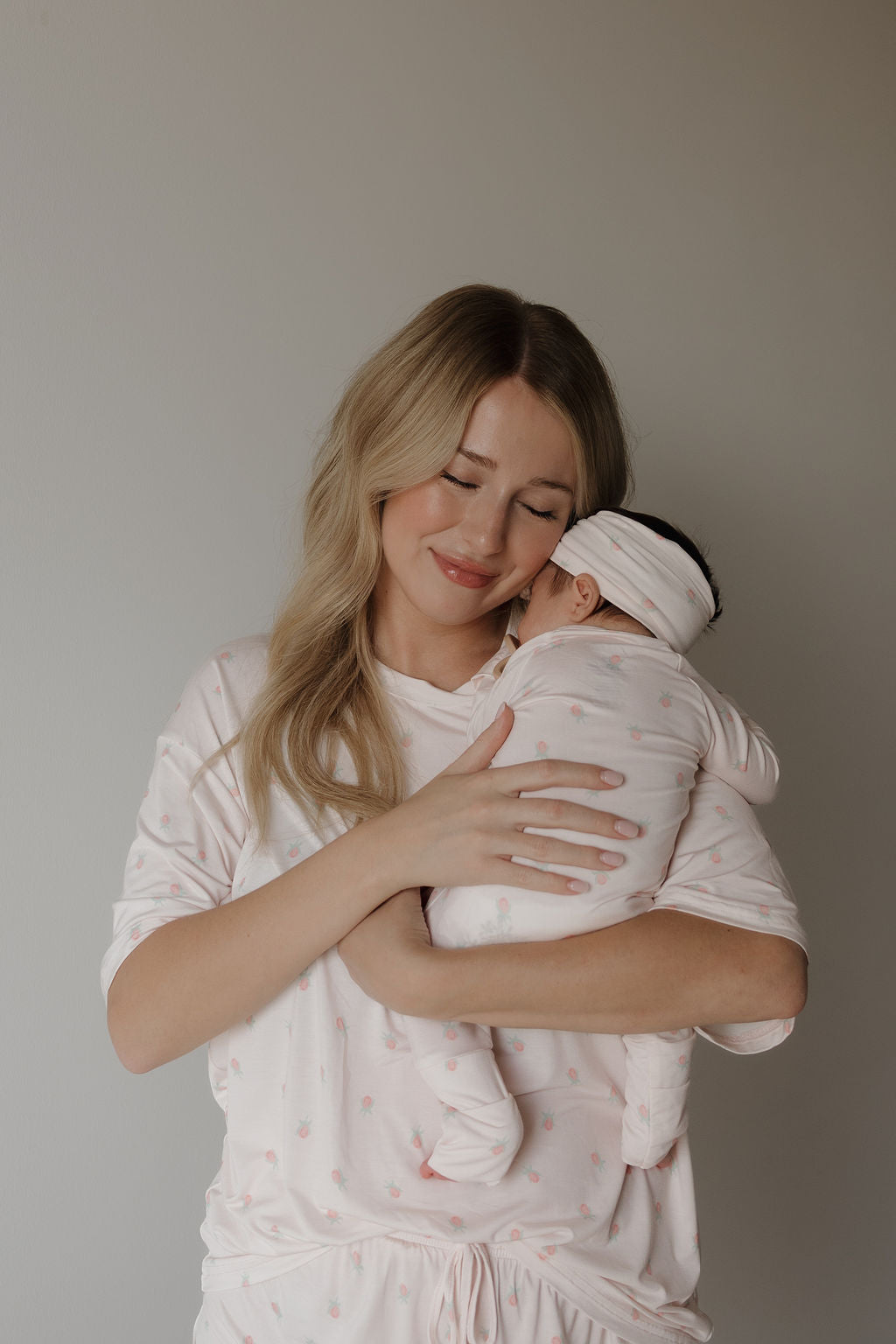 A woman with long blonde hair smiles softly while hugging a sleeping baby, both dressed in matching pale pink outfits from forever french baby, including the gentle and hypo-allergenic Women’s Bamboo Short Set | Rosie—made from breathable bamboo fabric that’s perfect for sensitive skin.