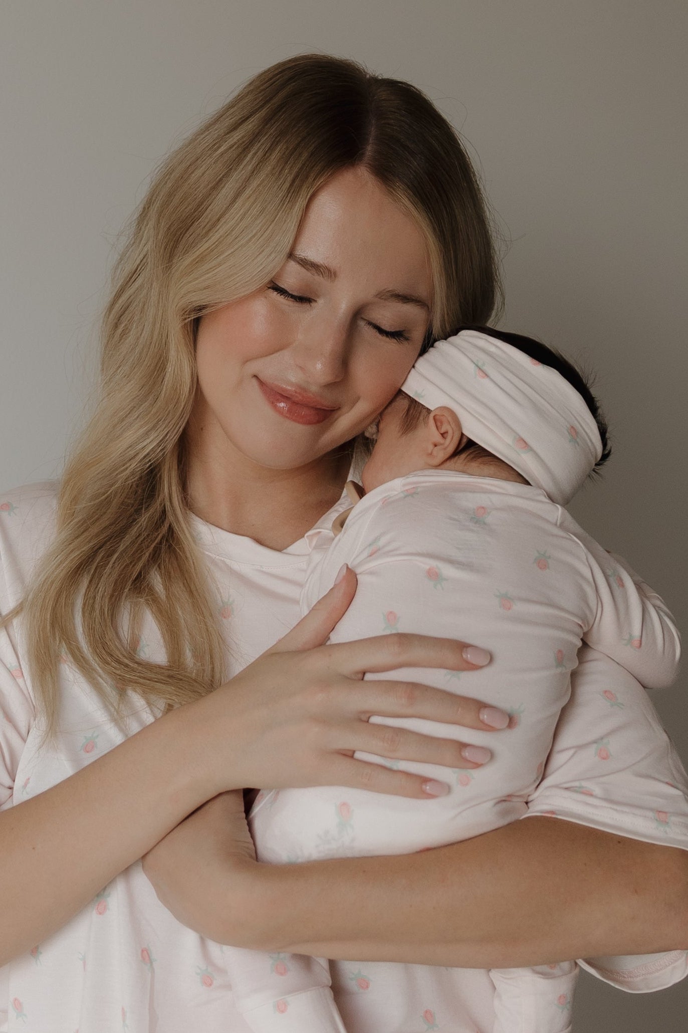 A woman with long blonde hair smiles softly while holding a baby dressed in matching light pink pajamas and the forever french baby Bamboo Head Wrap | Rosie with an adjustable bow. Resting comfortably on her shoulder against a plain background, the little one looks cozy and adorable in the bamboo head wrap.