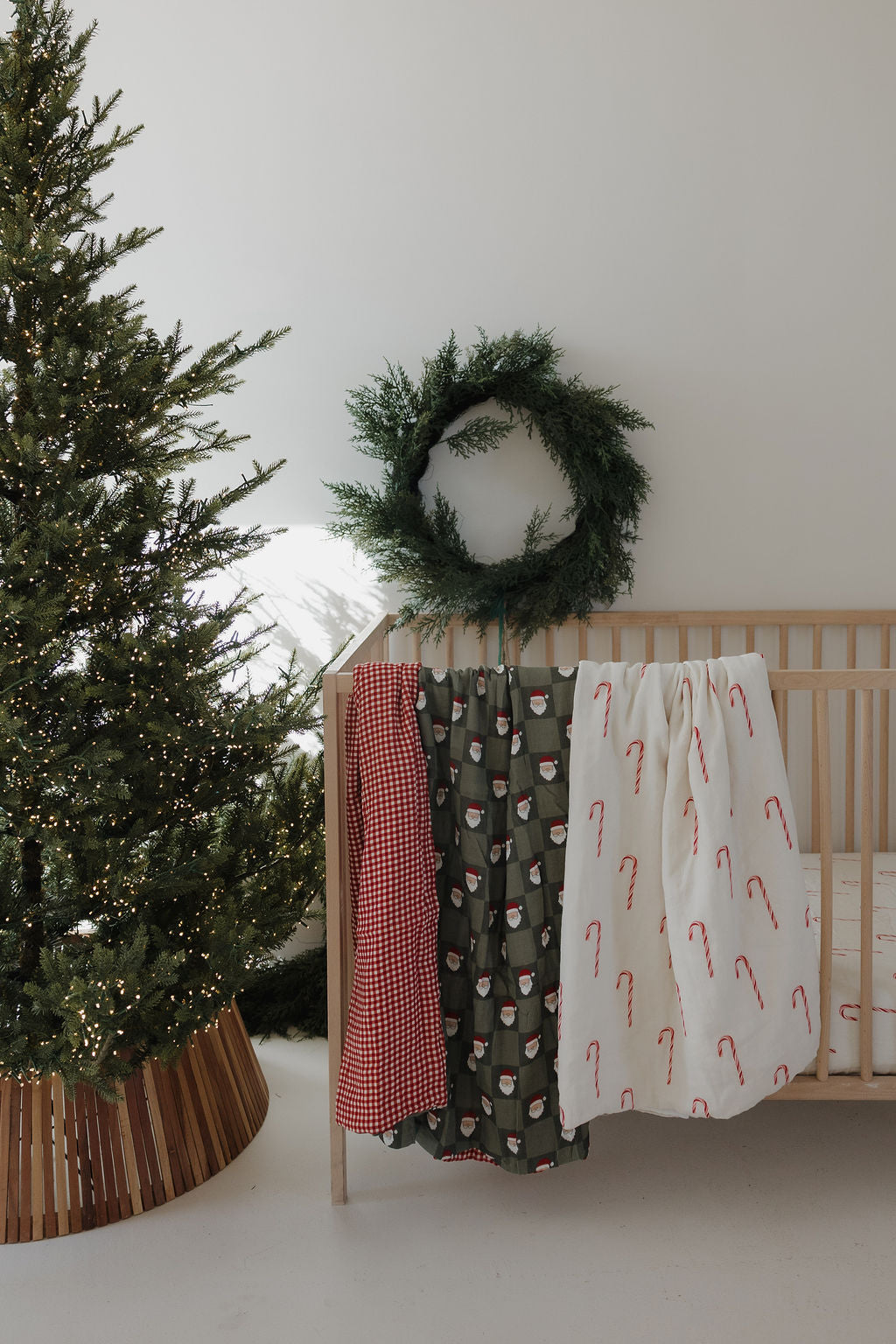 A decorated Christmas tree stands by a wooden crib dressed in the forever french baby Muslin Quilt | Red Gingham & Santa Checkerboard, with a green wreath above on a white wall.