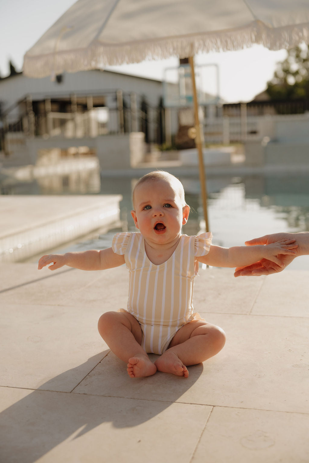 A baby in the lolo webb 2 Girl Sleeveless Swimsuit | Sun Rays with yellow-white stripes and ruffle detail sits poolside, arms outstretched, beneath a white umbrella as an adult's hand reaches toward her.