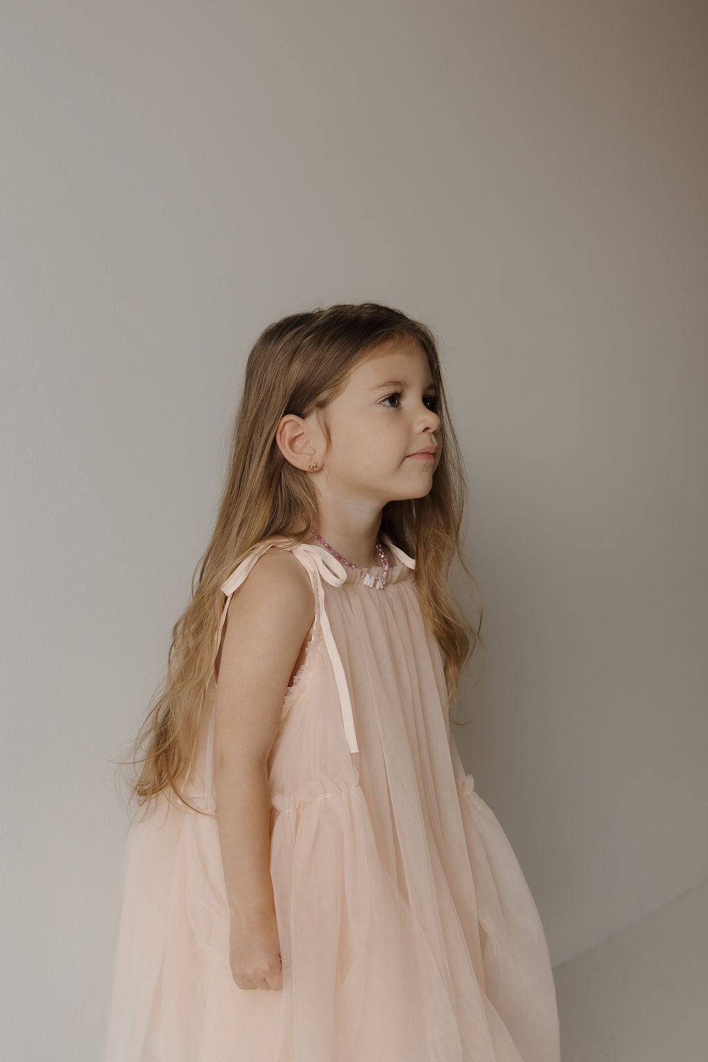 Wearing the forever french baby Tutu Dress | Slipper, a young girl with long brown hair stands against a plain, light-colored background and looks to the side. The dress is light pink and sleeveless with a tutu skirt.