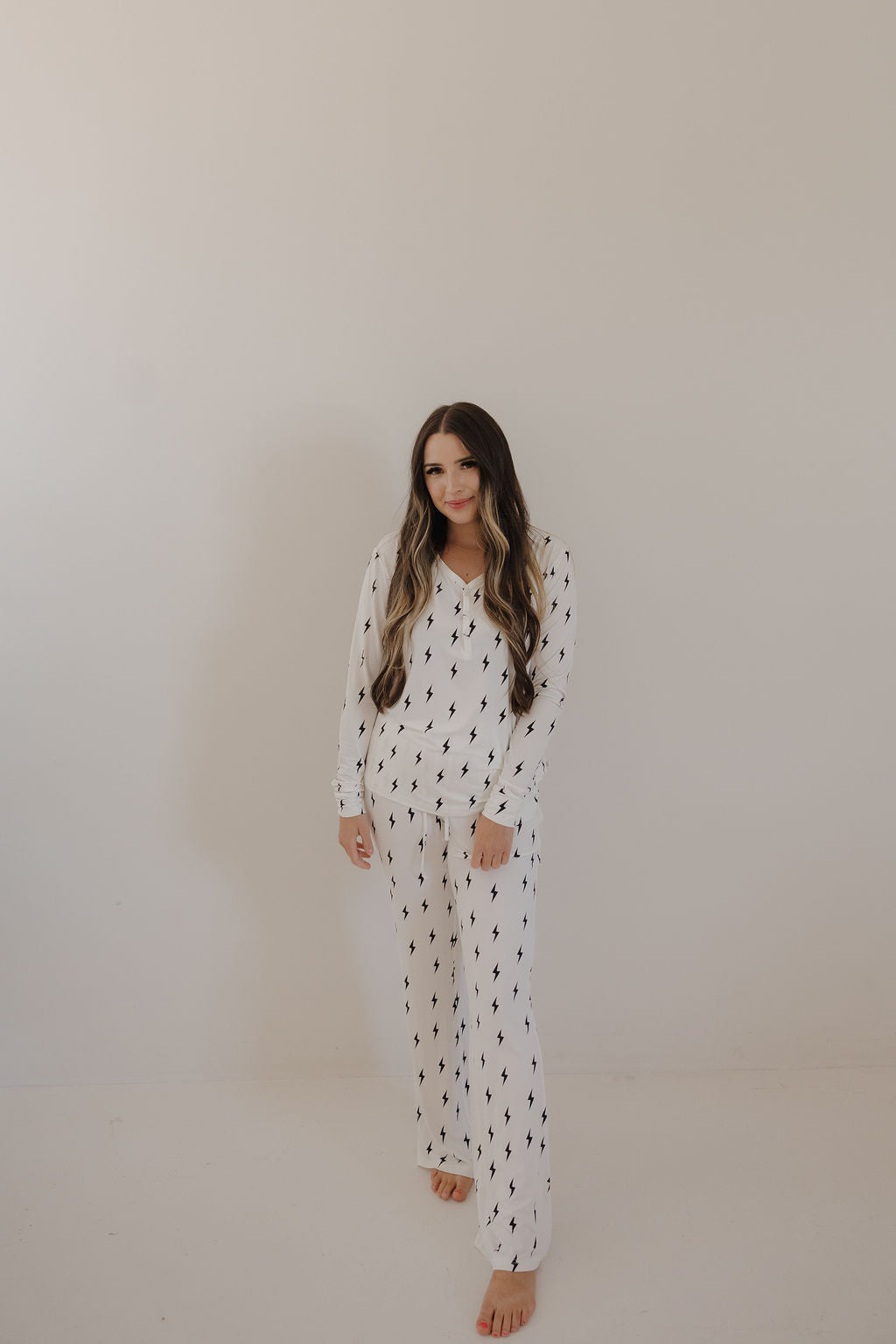 A woman with long, wavy hair stands barefoot against a white wall, softly smiling as she wears forever french baby's Women's Bamboo Pajama in White & Black Lightning Bolt—a breastfeeding-friendly set featuring a subtle black print.
