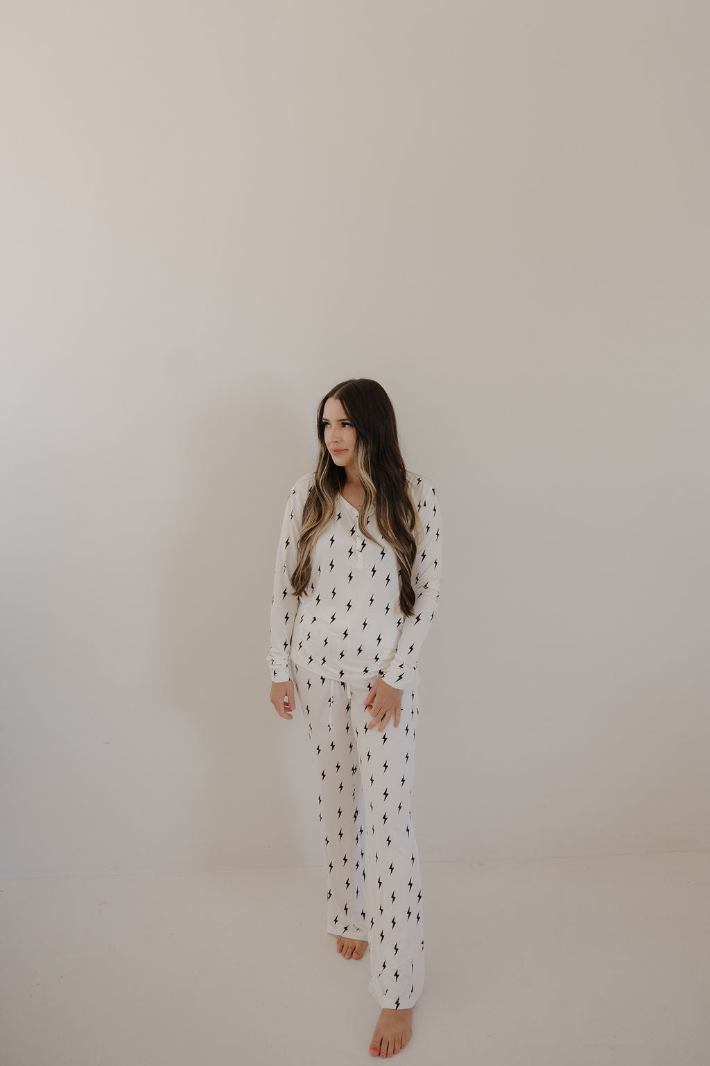 A woman with long brown hair stands barefoot against a plain light wall, wearing the forever french baby Women's Bamboo Pajama in White & Black Lightning Bolt. She gazes slightly to the side with a relaxed expression.