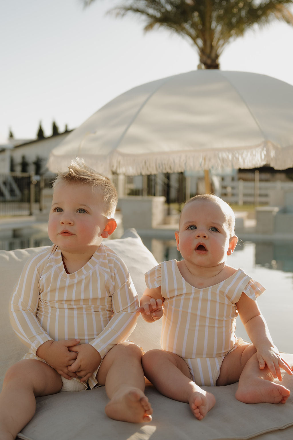 Two young children in matching lolo webb 2 Girl Sleeveless Swimsuit | Sun Rays, featuring yellow and white stripes with ruffle detail, sit by the pool under a large umbrella and palm tree, enjoying the warm sunlight.