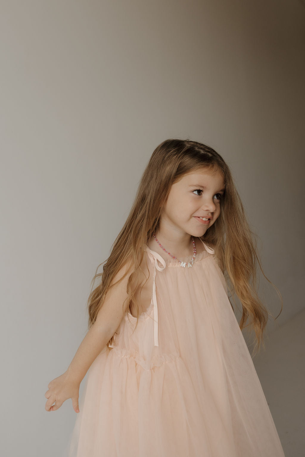 A young girl with long brown hair wears the forever french baby Tutu Dress | Slipper in light pink, featuring delicate bow straps and a beaded necklace, smiling and looking to the side against a neutral background.