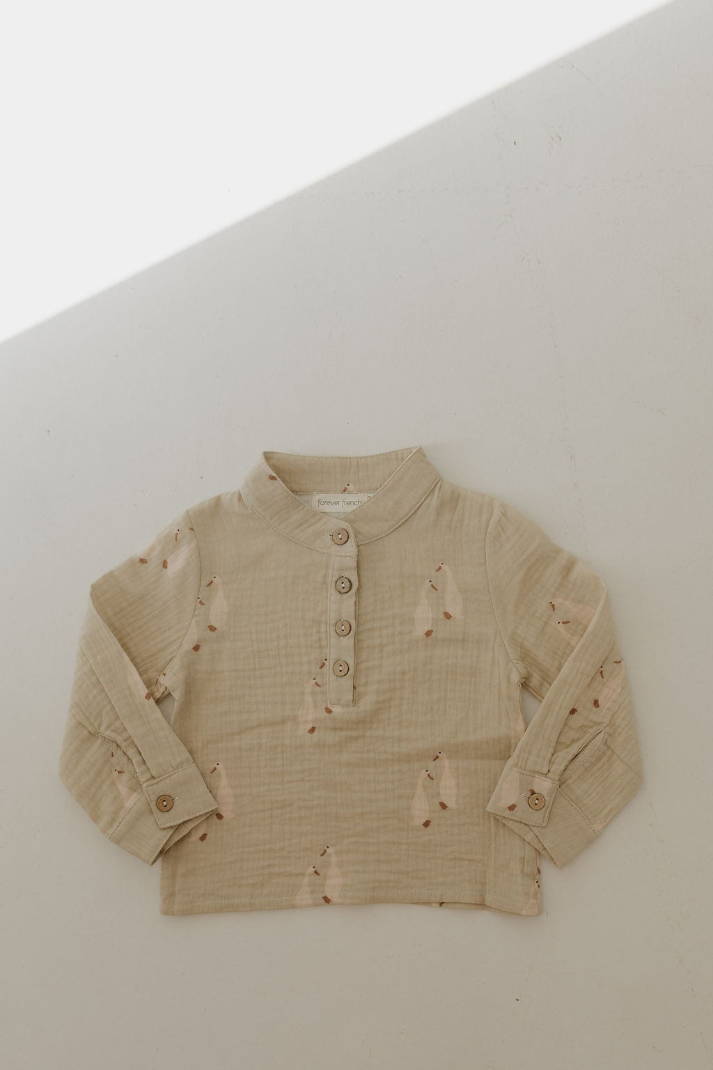 The Muslin Button Up | Silly Goose by forever french baby is a beige, long-sleeve, cotton shirt with a mandarin collar, wooden buttons, and subtle bunny print, shown laid flat on a light surface with a soft diagonal shadow.