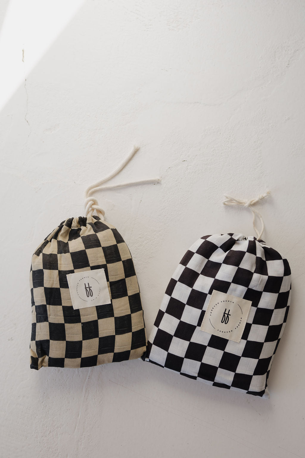 Two drawstring pouches featuring checkerboard patterns inspired by the Muslin Crib Sheet | Black & White Checkerboard from forever french baby, in beige/black and white/black, both with a minimalist logo label on a textured background.