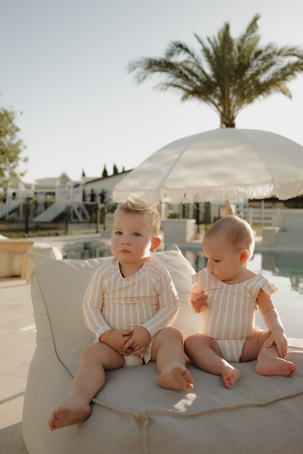 Two young children in matching lolo webb 2 Toddler Rash Guard | Sun Rays swimsuits relax on a lounge chair by the pool, enjoying a sunny day under a large umbrella and palm tree.