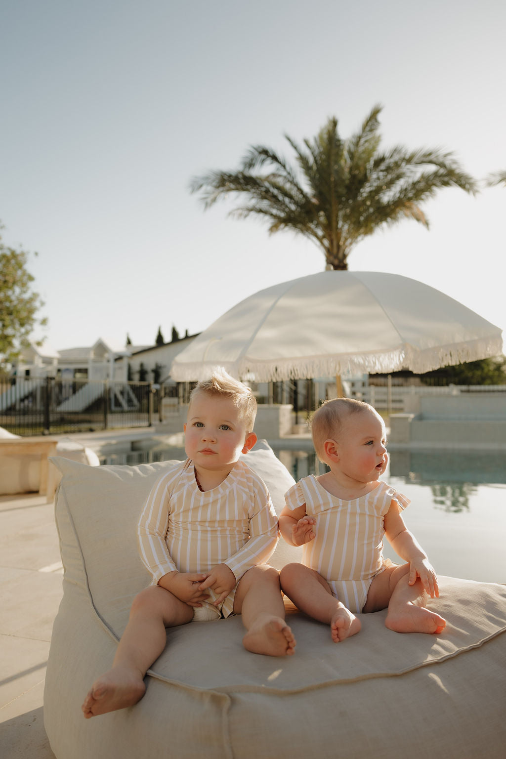 Two young children in matching lolo webb 2 Toddler Rash Guard | Sun Rays swimsuits sit on a beige outdoor lounger by the pool, with a large white umbrella and palm tree in the background on a sunny day.