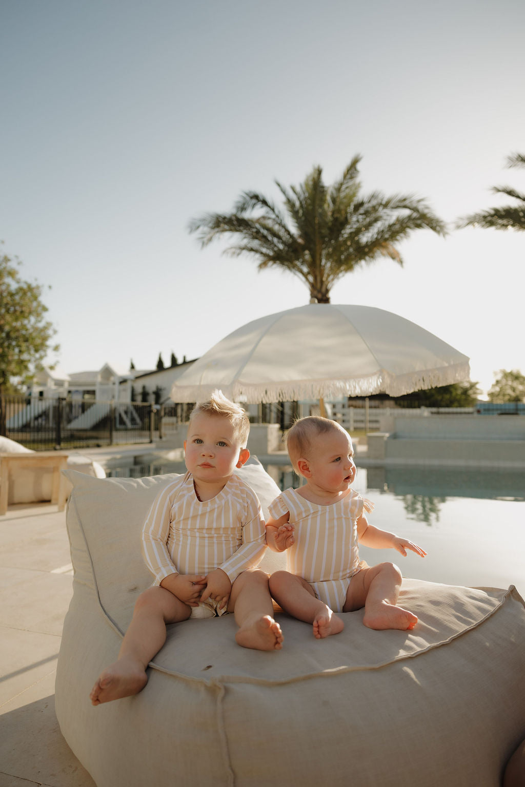 Two young children wearing lolo webb 2’s Toddler Rash Guard | Sun Rays sit together by a pool on a beanbag. Palm trees and a white umbrella create a sunny, relaxed vibe in this perfect summer scene.