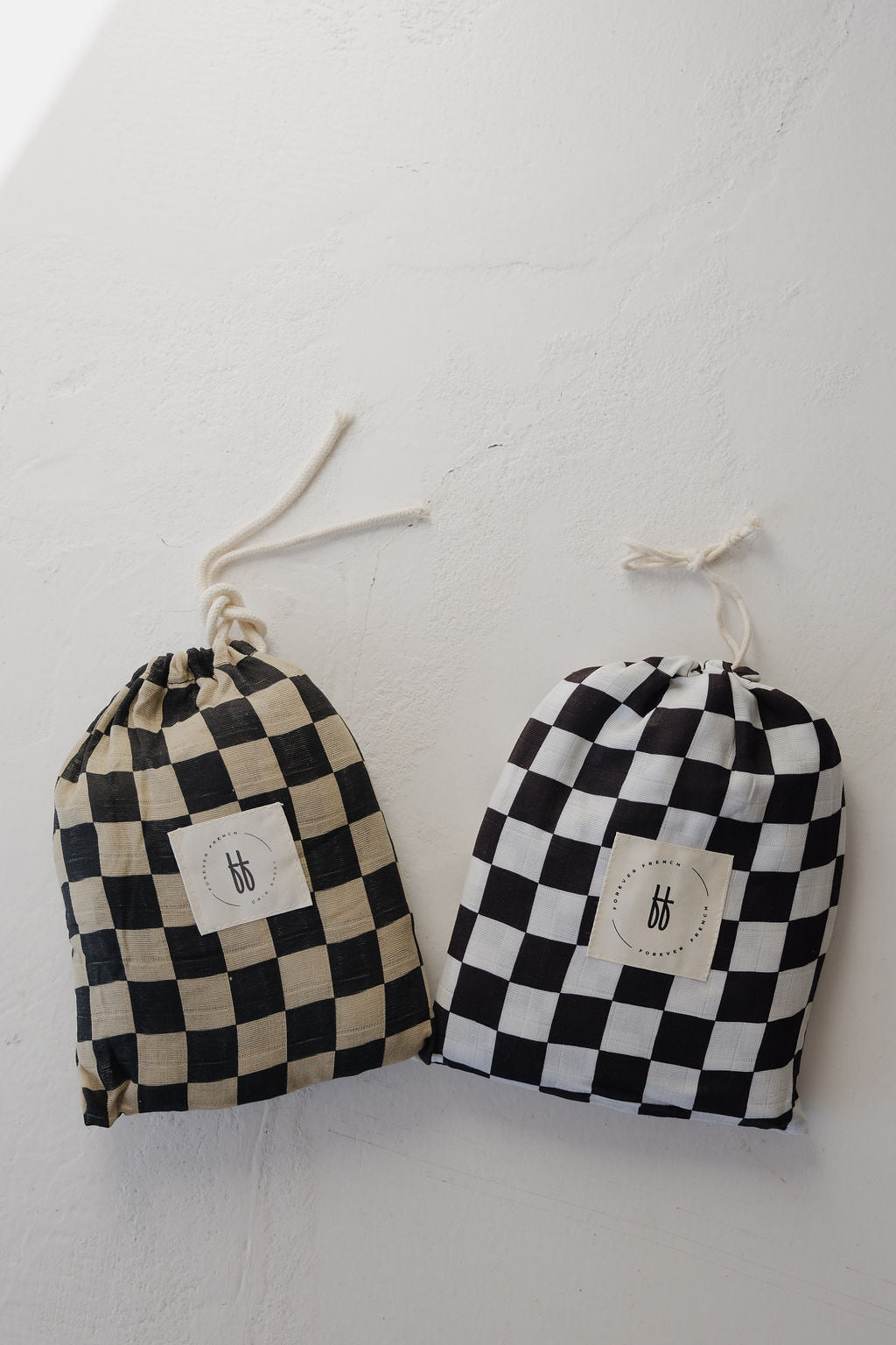 Two drawstring bags with beige-black and white-black checkered patterns, each featuring a circular logo label, rest on a white surface. Their design is inspired by the forever french baby Muslin Crib Sheet | Black Checkerboard.