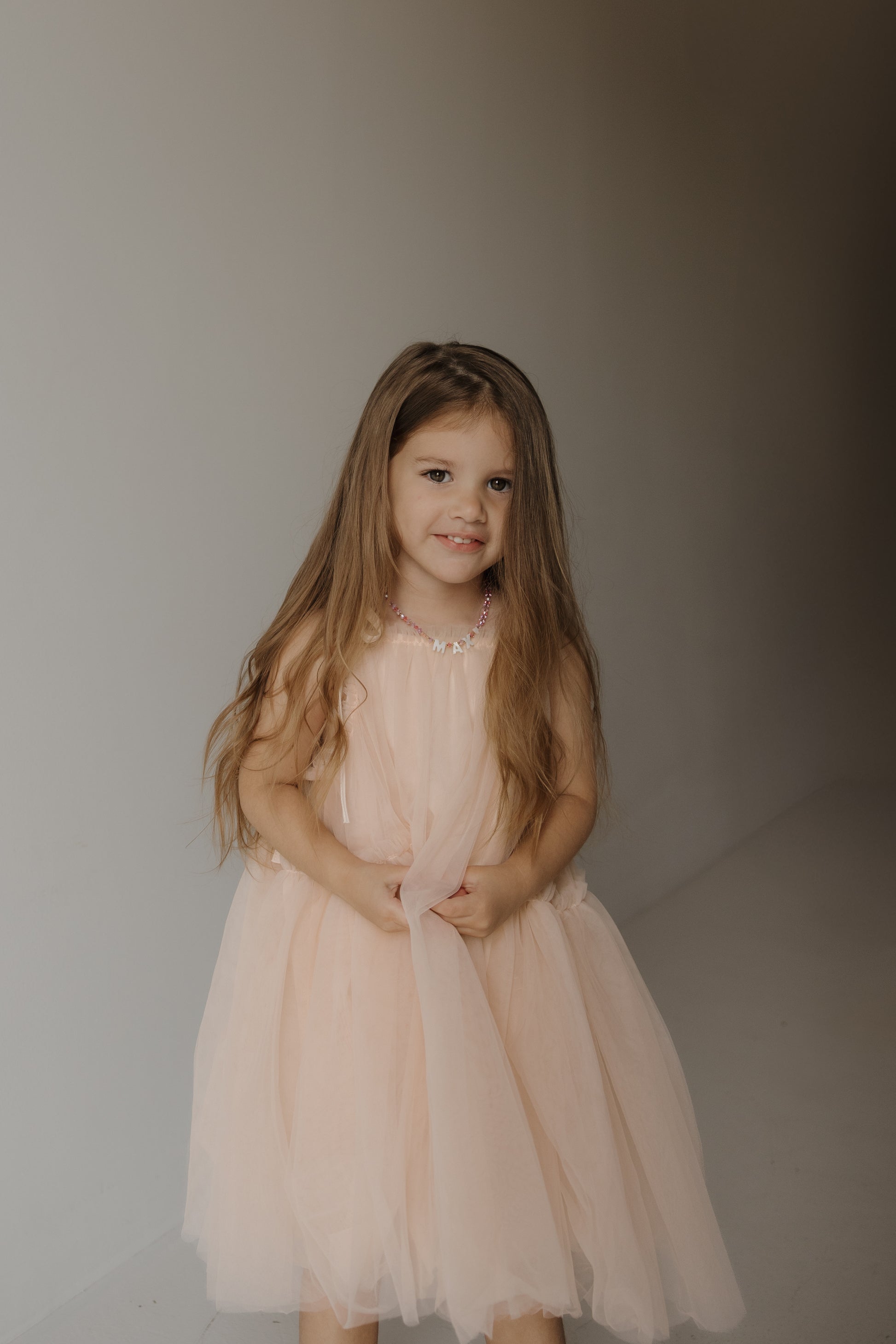 A young girl with long brown hair stands indoors against a neutral background, smiling softly at the camera while holding the skirt of her forever french baby Tutu Dress | Slipper, featuring delicate bow straps and a light pink tulle design.