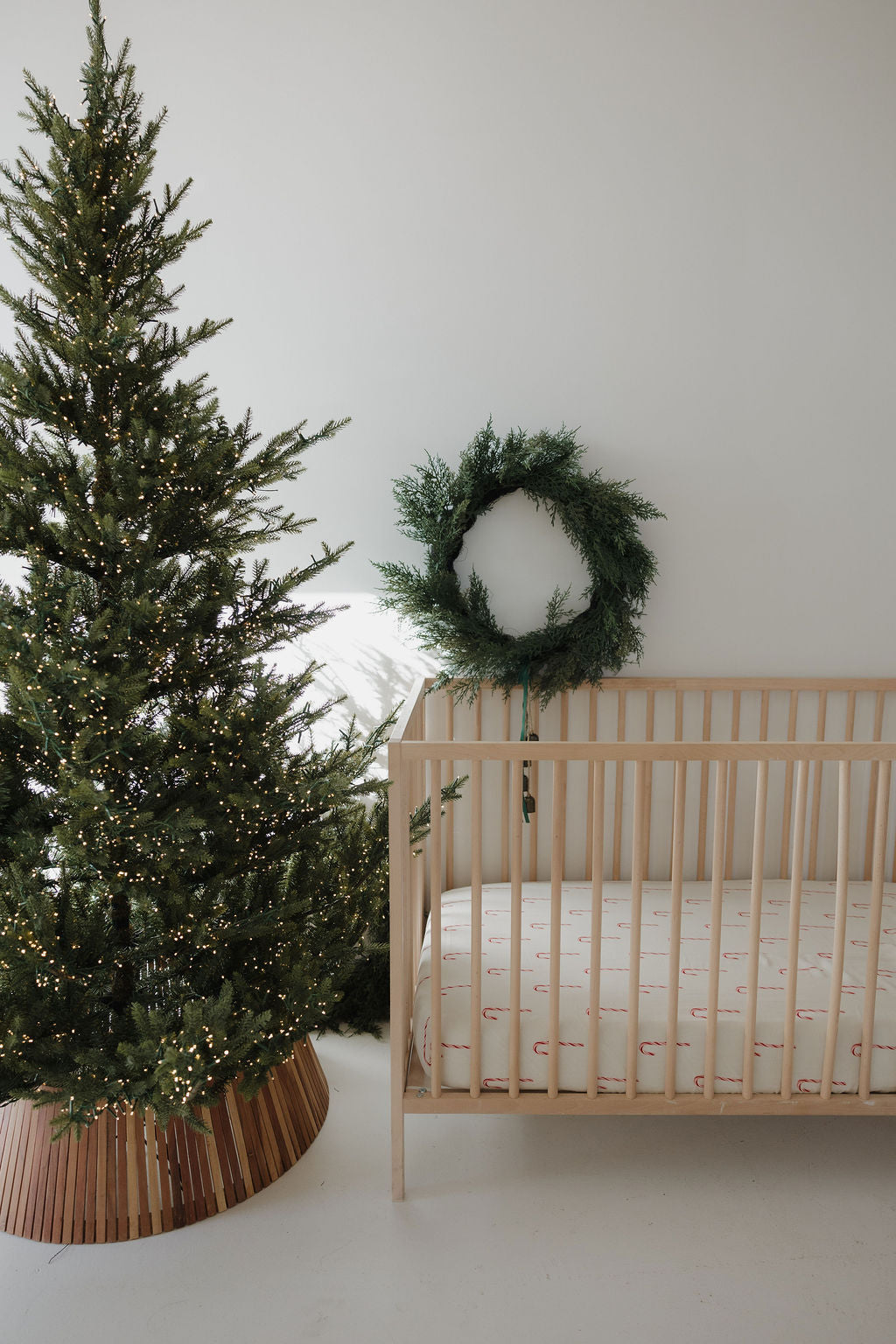 A minimal nursery featuring the forever french baby Muslin Crib Sheet | Candy Cane, a lit Christmas tree, and a green wreath on a white wall creates a festive and cozy atmosphere.