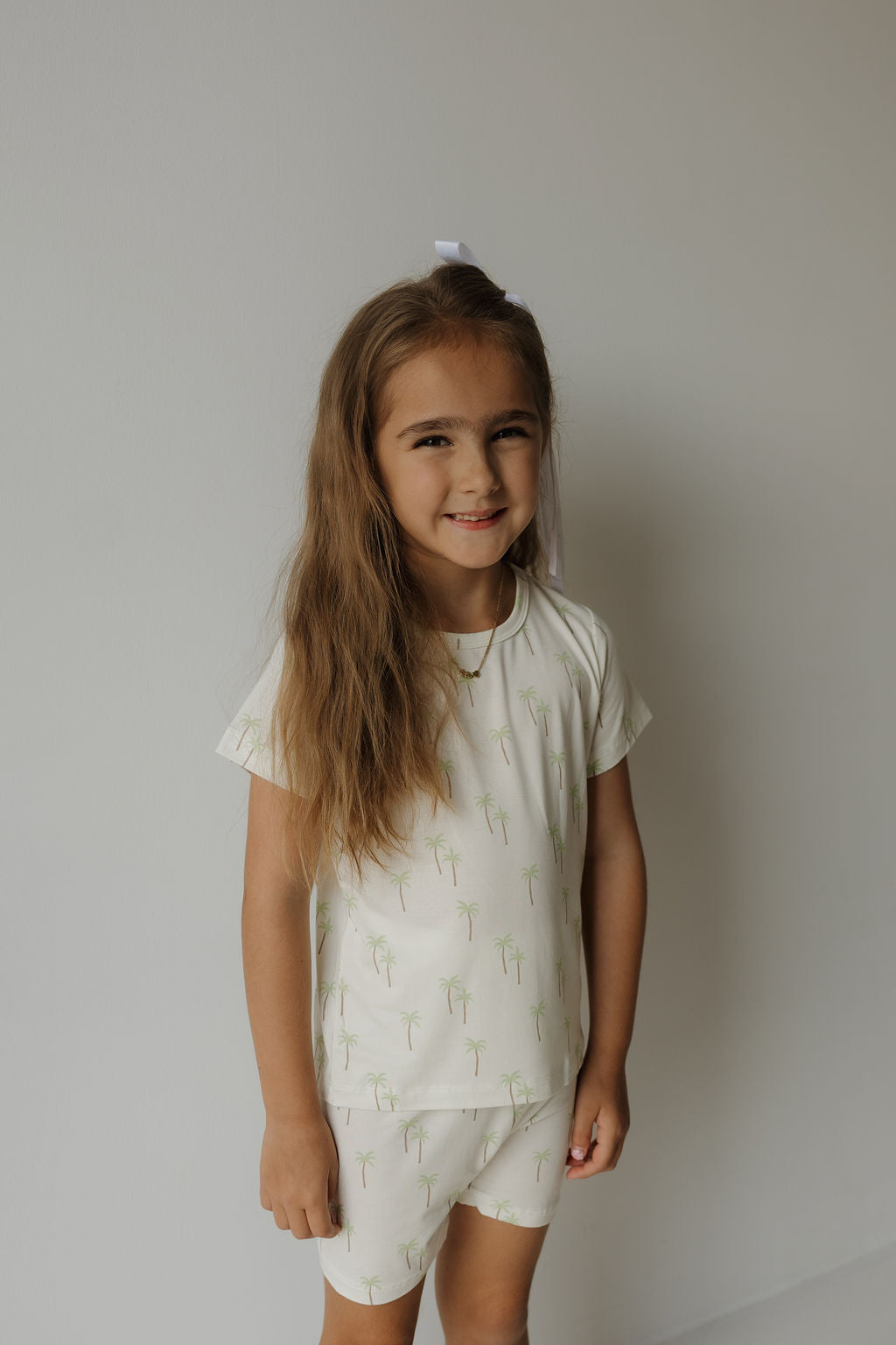 A young girl with long brown hair and a white bow smiles in front of a plain light-colored wall, wearing the Bamboo Short Set | Palm by forever french baby—a cream-colored t-shirt and shorts set featuring a subtle palm tree print, crafted from ultra-soft bamboo fabric for hypoallergenic comfort, ideal for kids’ sleepwear.