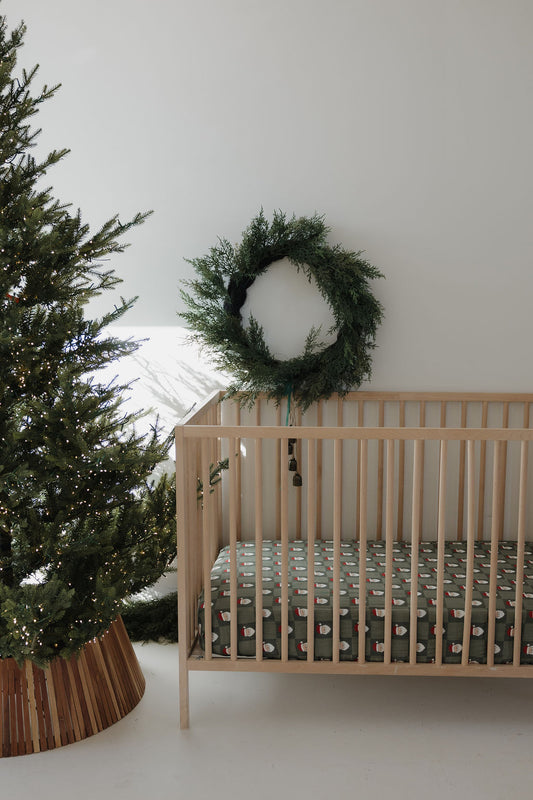 A simple nursery with a wooden crib featuring the forever french baby Muslin Crib Sheet | Santa Checkerboard, festive bedding, a large green wreath on the wall, and a decorated Christmas tree with lights on the left.