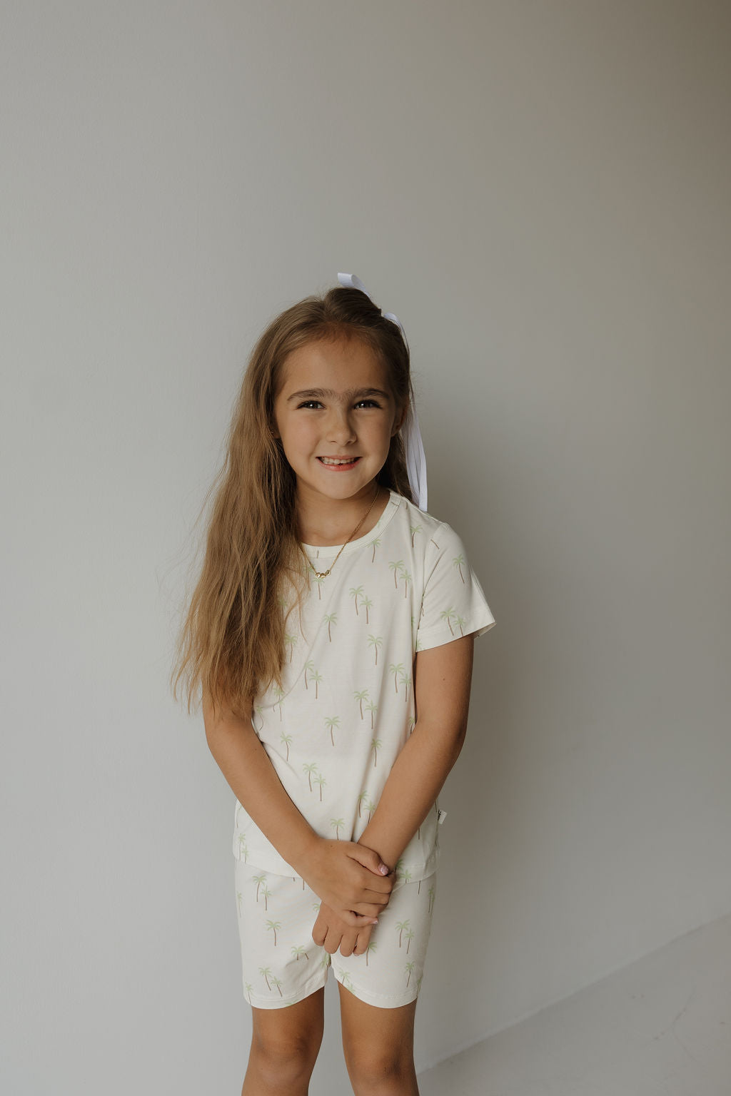 A young girl with long brown hair, wearing the forever french baby Bamboo Short Set | Palm—a white bamboo pajama outfit with small palm tree prints—stands against a plain light background, smiling with her hands clasped in front of her.