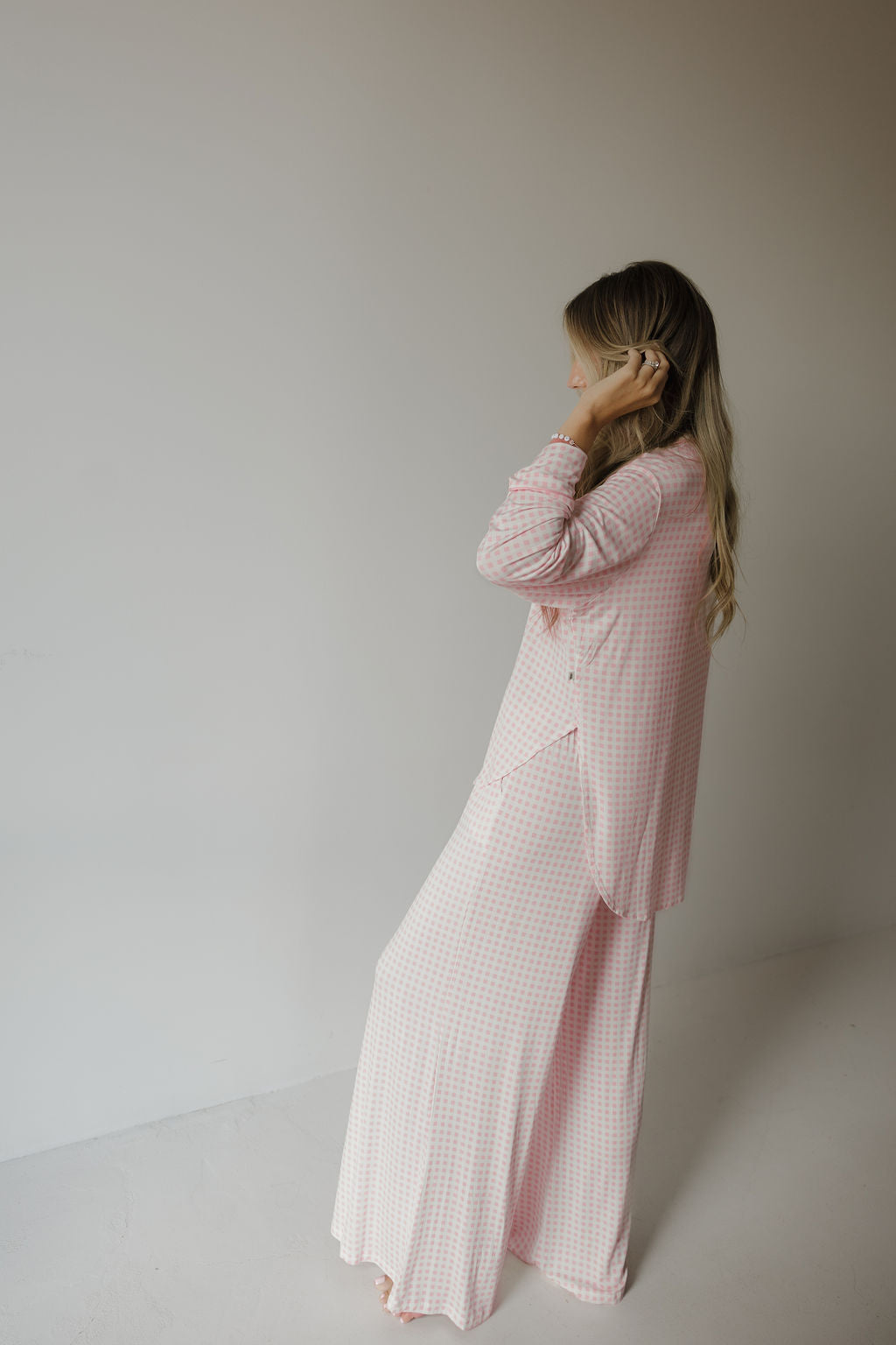 A person with long hair stands in a corner, wearing the Giselle Women’s Bamboo Button Up Lounge Set by forever french baby. They are dressed in a light pink and white checkered set from the women’s bamboo clothing collection, facing sideways and touching their hair against a neutral background.
