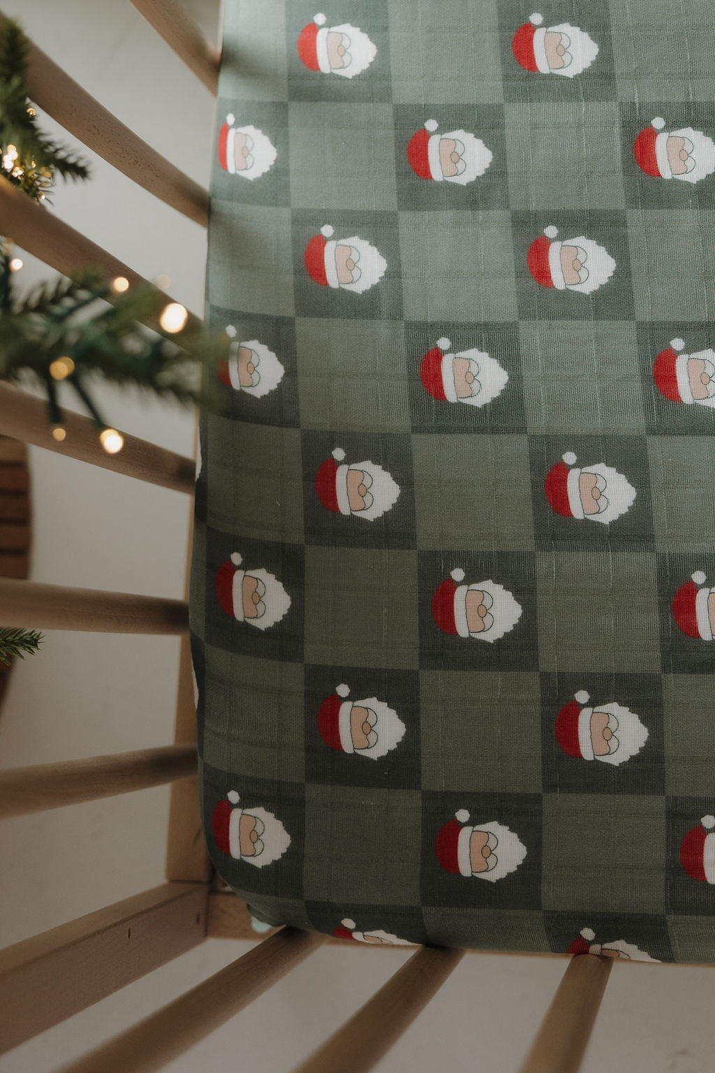 The Muslin Crib Sheet | Santa Checkerboard (Preview) by forever french baby features soft muslin cotton with Santa faces in a green checkered pattern, shown from above in a wooden crib accented by Christmas tree branches.