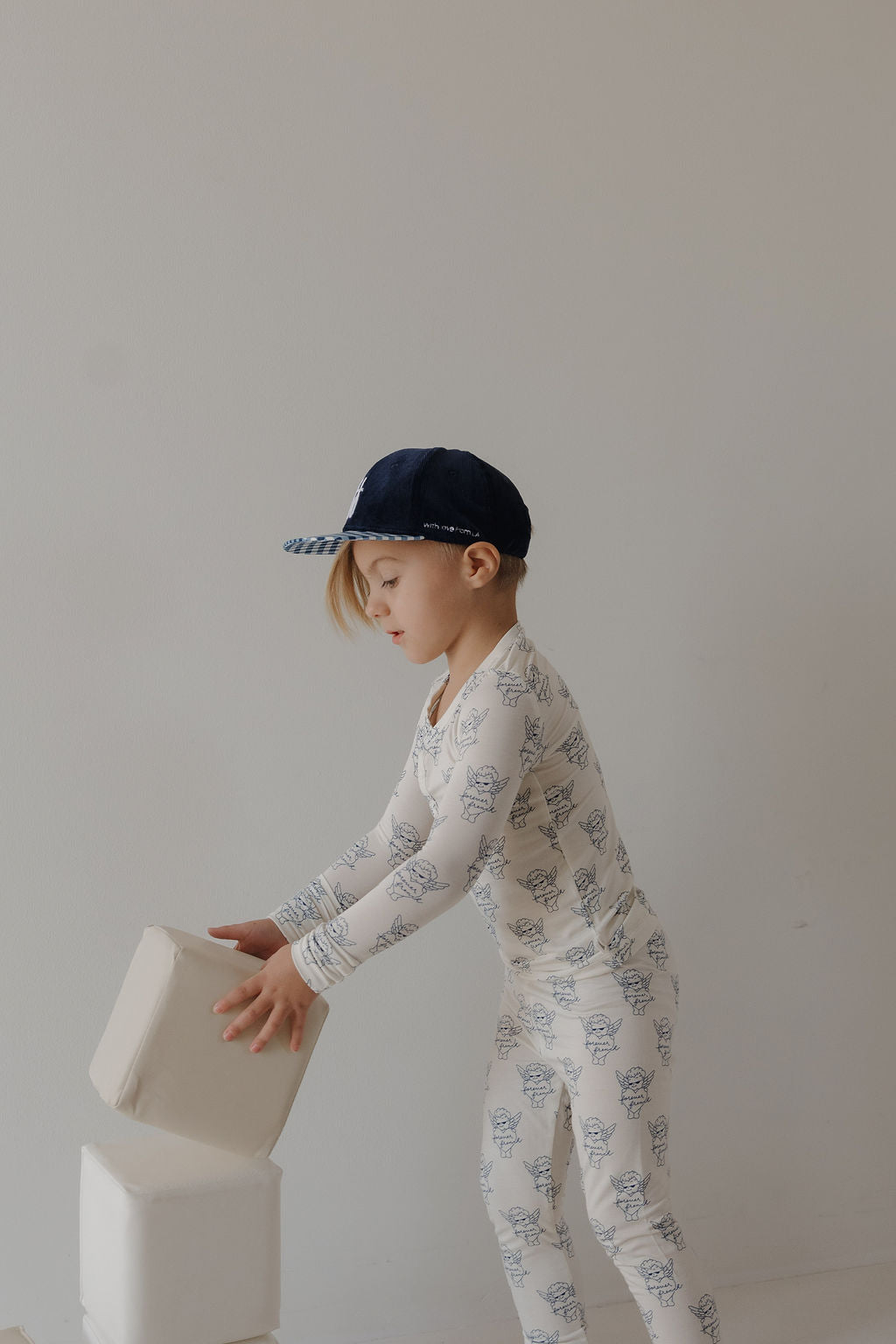 A young child in forever french baby Bamboo Two Piece Pajamas | ff Los Angeles and a navy blue cap stacks large white foam blocks against a plain light-colored wall.