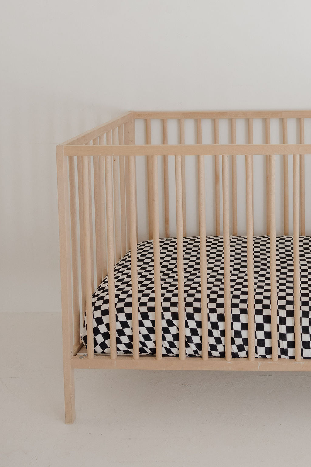 A light wooden crib with vertical slats holds the forever french baby Muslin Crib Sheet | Black & White Checkerboard, set against a plain white wall and floor—perfect for stylish baby bedding.