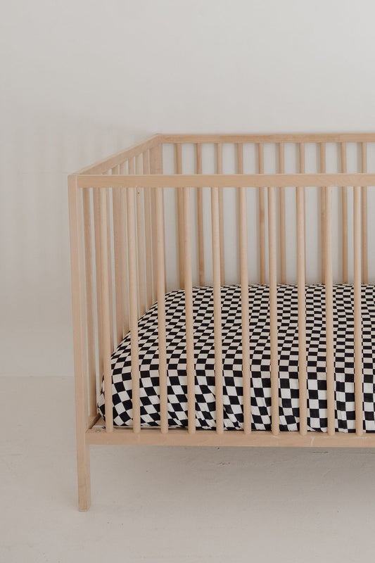 A light wooden crib with vertical slats holds the forever french baby Muslin Crib Sheet | Black & White Checkerboard, set against a plain white wall and floor—perfect for stylish baby bedding.