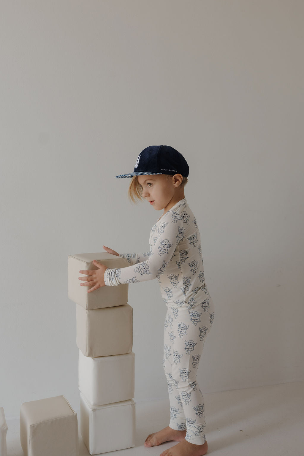 A young child in forever french baby’s Bamboo Two Piece Pajamas by ff Los Angeles and a navy cap stands barefoot, stacking large white blocks against a plain light wall.