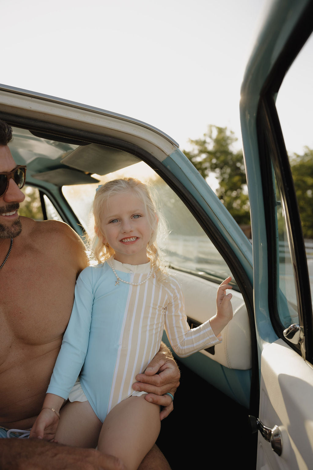 A young girl in the lolo webb 2 Girl Long Sleeve Swimsuit | Surf & Sand sits on a man's lap in a sunlit car with the door open, smiling at the camera, with trees in the background.