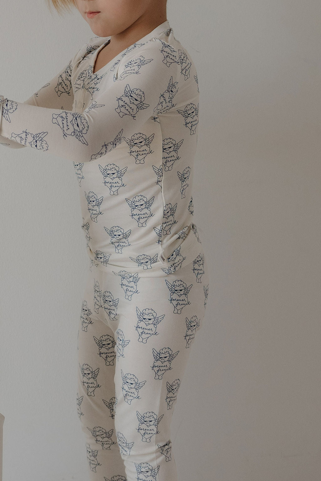 A young child wears forever french baby’s Bamboo Two Piece Pajamas from ff Los Angeles, featuring a cream color with blue cherub print. The stylish, breathable sleepwear stands out against a plain light background.