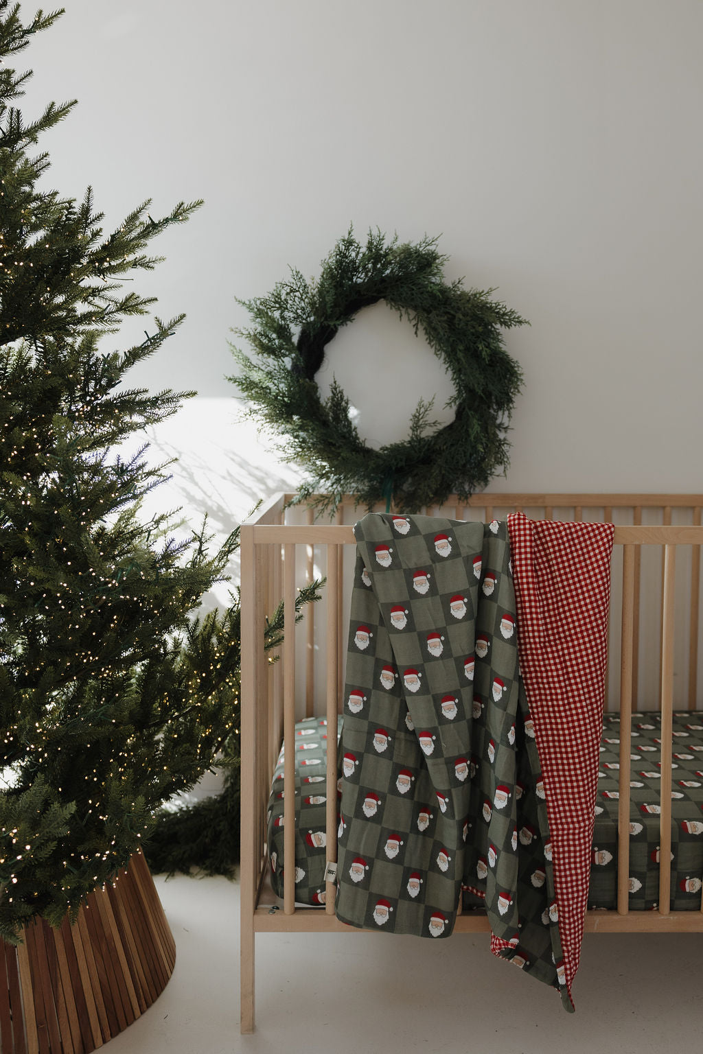 A wooden crib features the forever french baby Muslin Quilt | Red Gingham & Santa Checkerboard (Preview), accented by a green crib sheet with Santa faces, a hanging green wreath, and a nearby Christmas tree with twinkling lights.