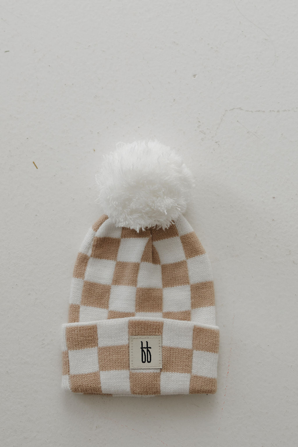 The forever french baby ff Knit Pom Top Beanie | Tan Checkerboard features a beige and white checkerboard knit, fluffy white pom-pom, folded brim with a black double "b" logo patch, and is shown laid flat on a light surface.