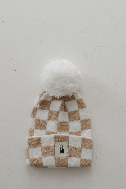 The forever french baby ff Knit Pom Top Beanie | Tan Checkerboard features a beige and white checkerboard knit, fluffy white pom-pom, folded brim with a black double "b" logo patch, and is shown laid flat on a light surface.