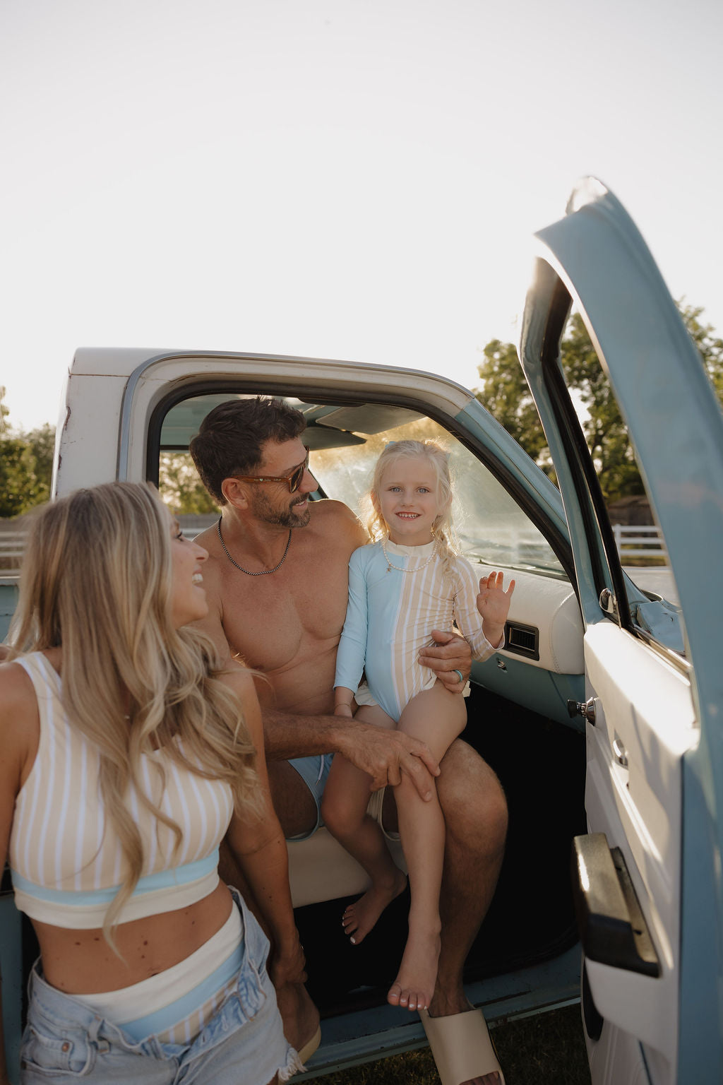A man sits in the open door of a vintage blue truck, holding a smiling young girl wearing the lolo webb 2 Girl Long Sleeve Swimsuit | Surf & Sand. Nearby, a woman smiles as they all enjoy a sunny outdoor moment together.