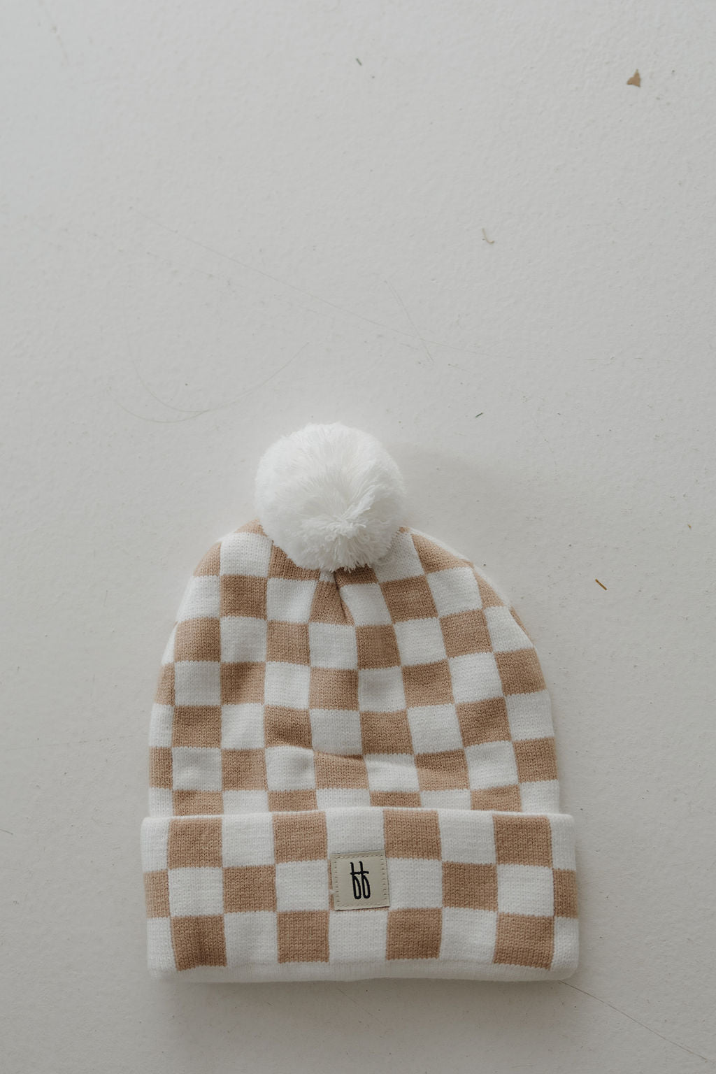The forever french baby Adult ff Knit Pom Top Beanie in Tan Checkerboard features a white and light brown checkerboard pattern, ribbed folded brim with small logo, and fluffy white pom-pom on top, shown laid flat on a white surface.