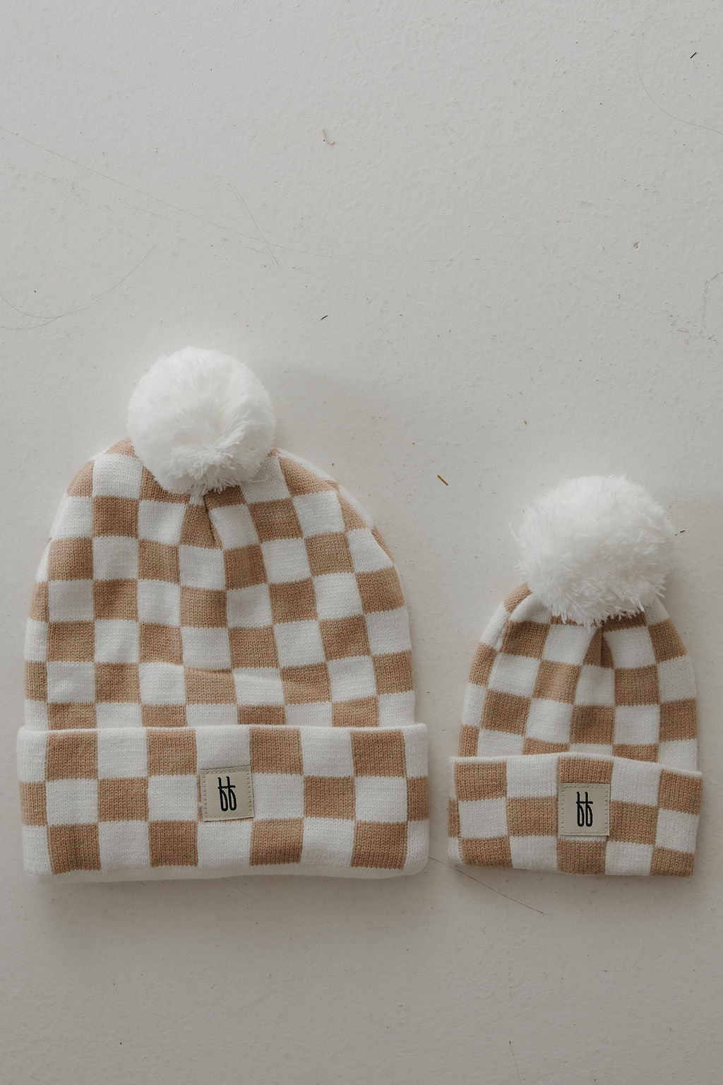 Two ff Knit Pom Top Beanies in Tan Checkerboard by forever french baby—one adult and one baby size—are side by side on a light surface, each with a "bb" logo label on the folded brim and a white pom-pom on top.