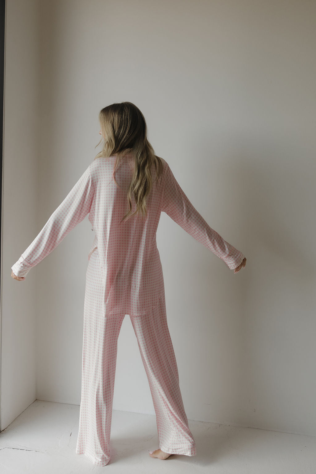 A woman with long hair stands barefoot facing a white wall, arms slightly outstretched, wearing the forever french baby Women’s Bamboo Button Up Lounge Set | Giselle in a loose pink and white checkered pattern.