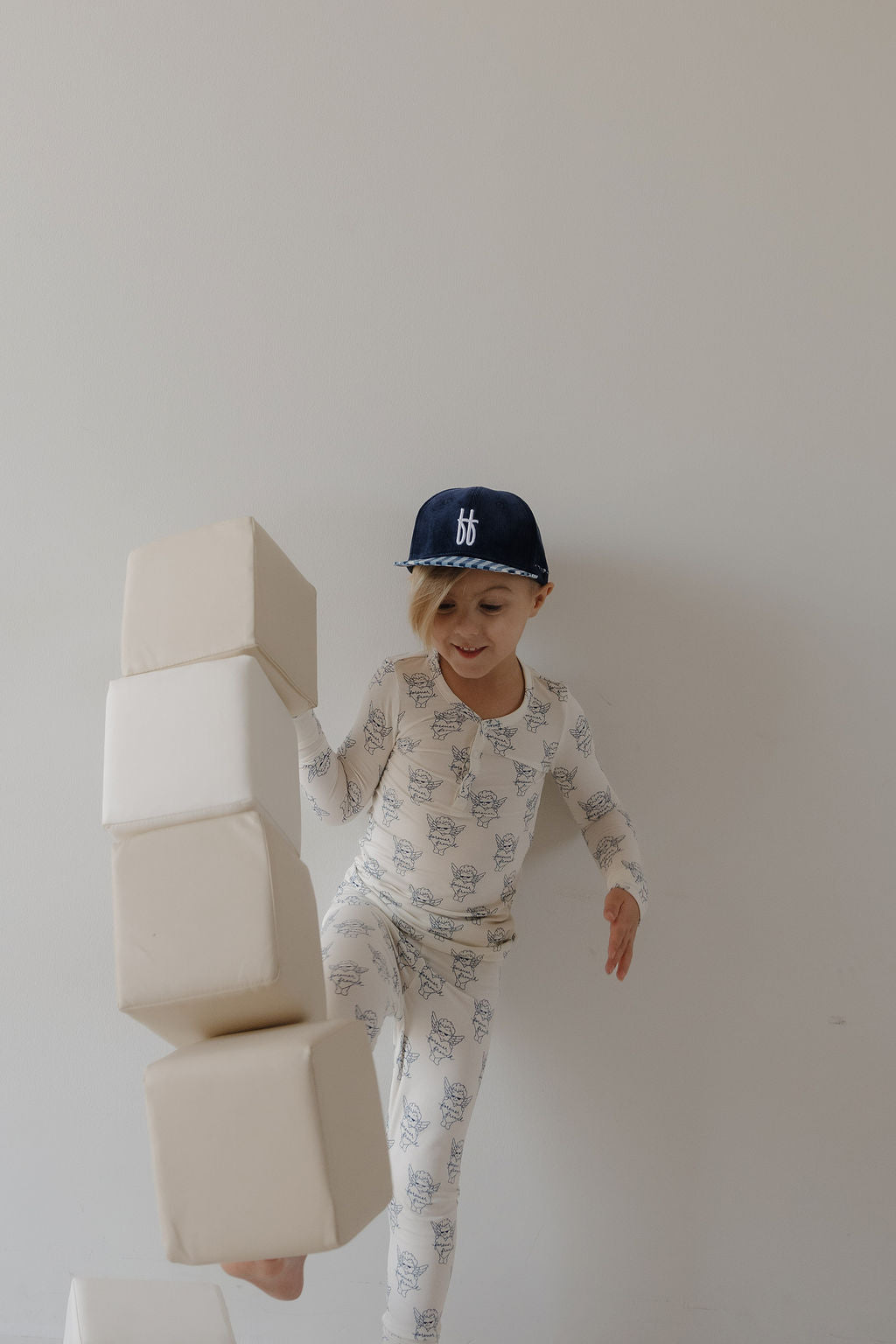 A young child in forever french baby Bamboo Two Piece Pajamas by ff Los Angeles and a navy cap kicks over large soft white blocks, smiling against a plain light background.