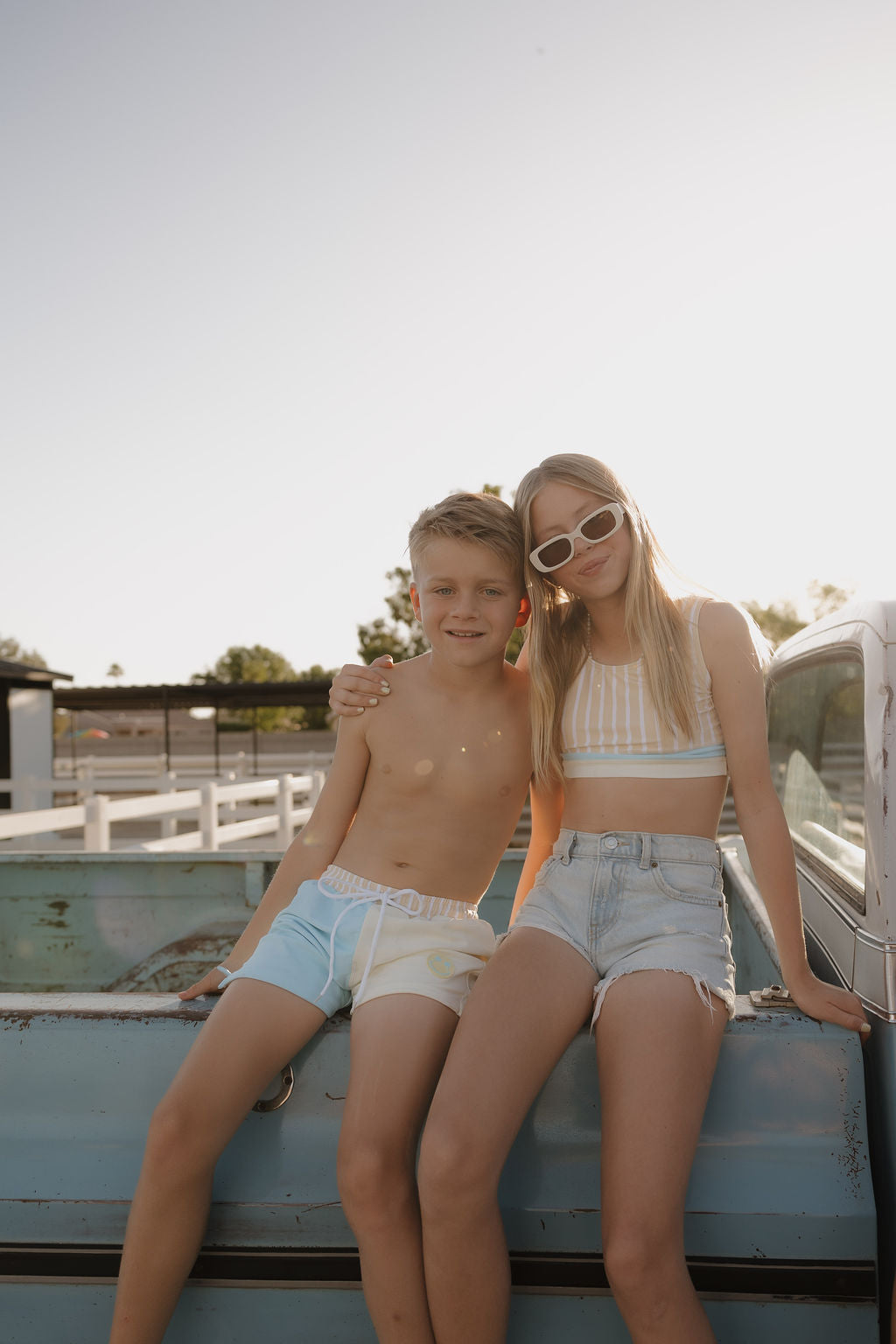Two kids sit on a pickup truck's tailgate. The boy wears lolo webb 2 Child Board Short | Surf & Sand, and the girl sports a striped crop top and denim shorts from our limited edition collection, smiling in the sunlight.