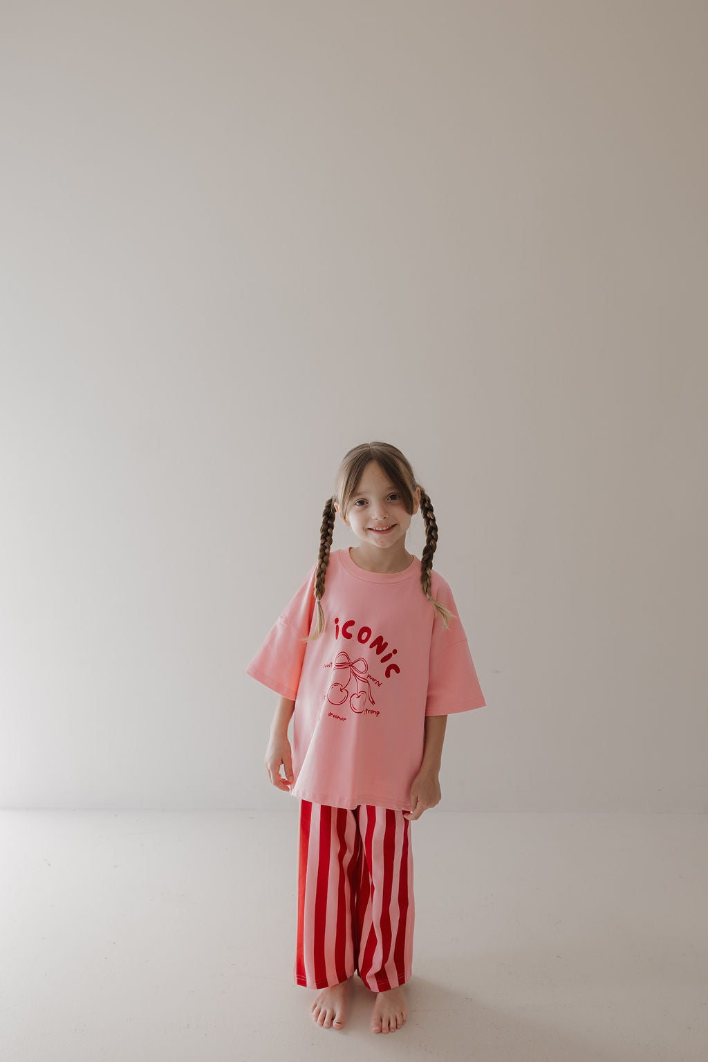 A young girl smiles softly at the camera, barefoot in a minimal room, wearing the Minty Cherries T-Shirt & Pant Set | Iconic Cherry & Stripe—a pink top with "ICONIC" and cartoon graphic, paired with loose red-and-white striped pants. Playful kids' fashion!.