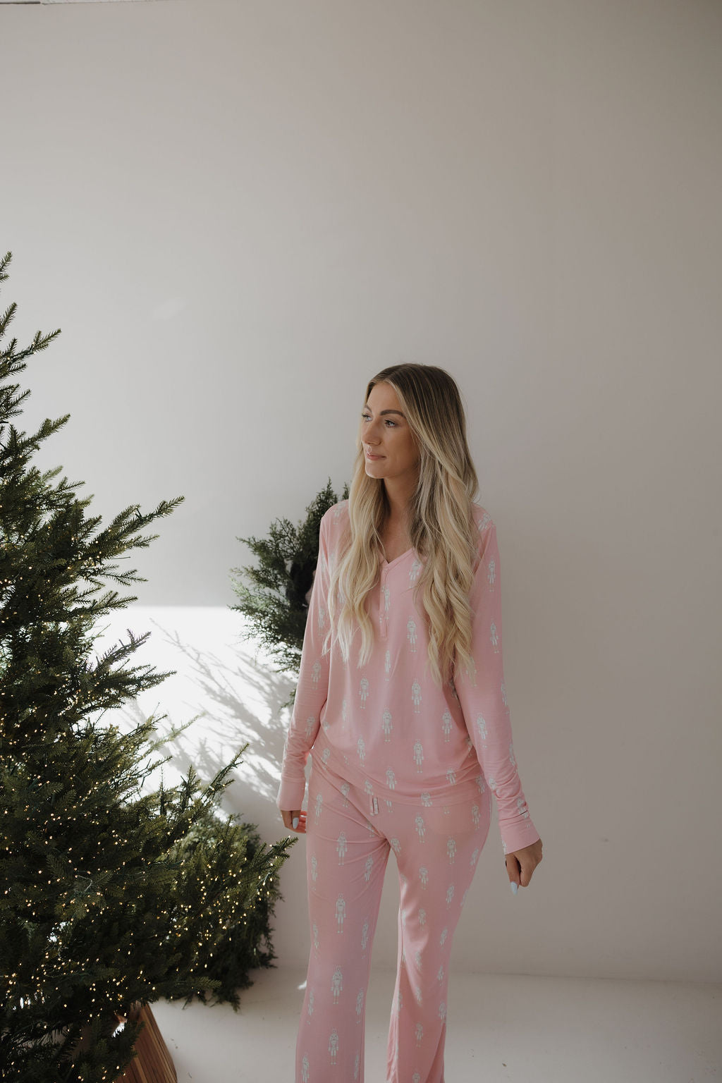 A woman with long blonde hair stands indoors near a decorated Christmas tree, wearing forever french baby Women's Bamboo Pajamas | Nutcracker, featuring a light pink, breastfeeding-friendly design with a small blue pattern.
