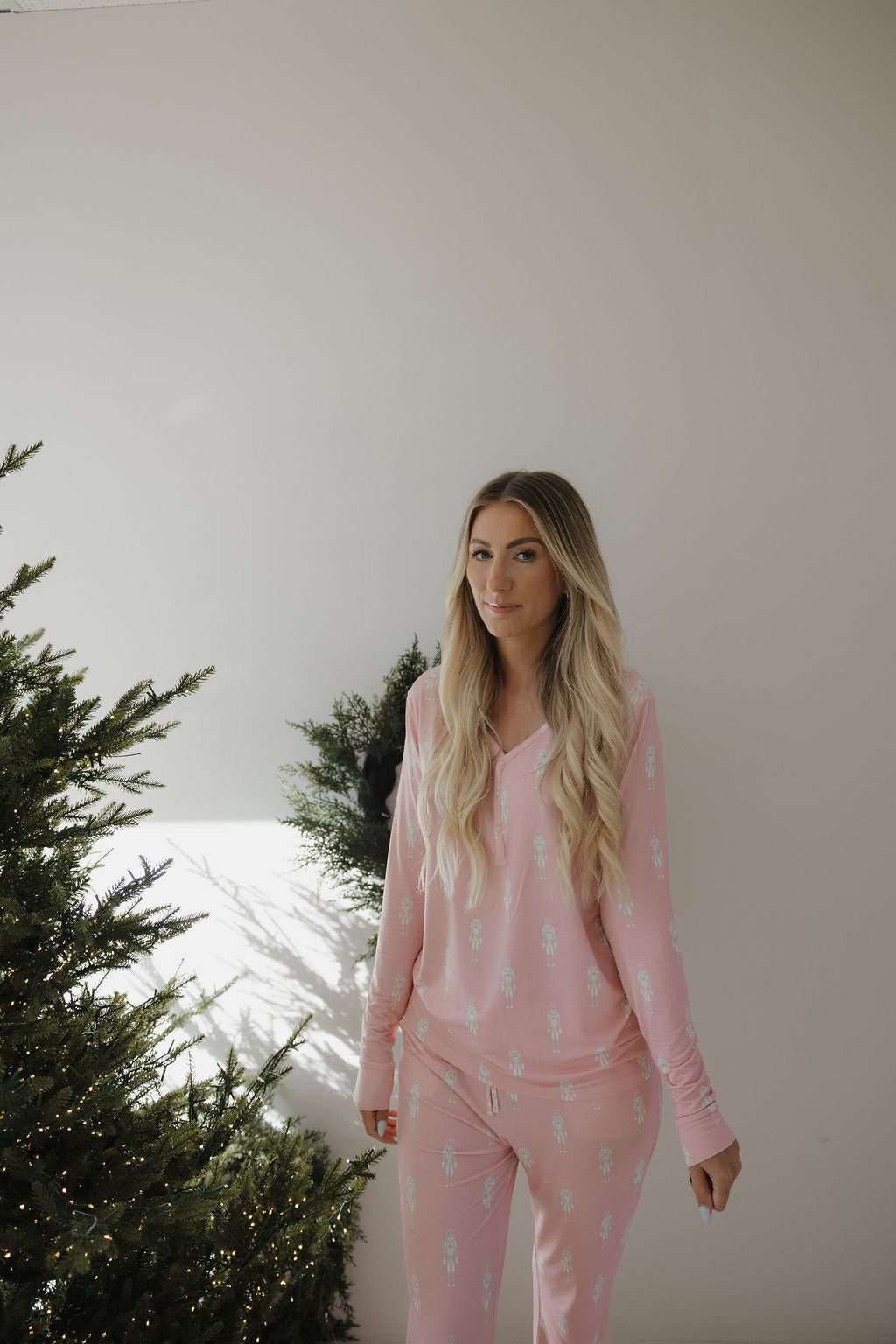 A woman with long blonde hair stands indoors beside a Christmas tree, wearing forever french baby Women's Bamboo Pajamas | Nutcracker. The minimal background features a plain white wall and touches of greenery.
