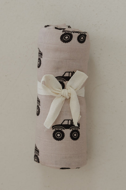 The forever french baby Muslin Swaddle | Monster Truck features ultra-soft beige cotton with black monster truck prints, rolled up and tied with a white ribbon, displayed on a light surface.